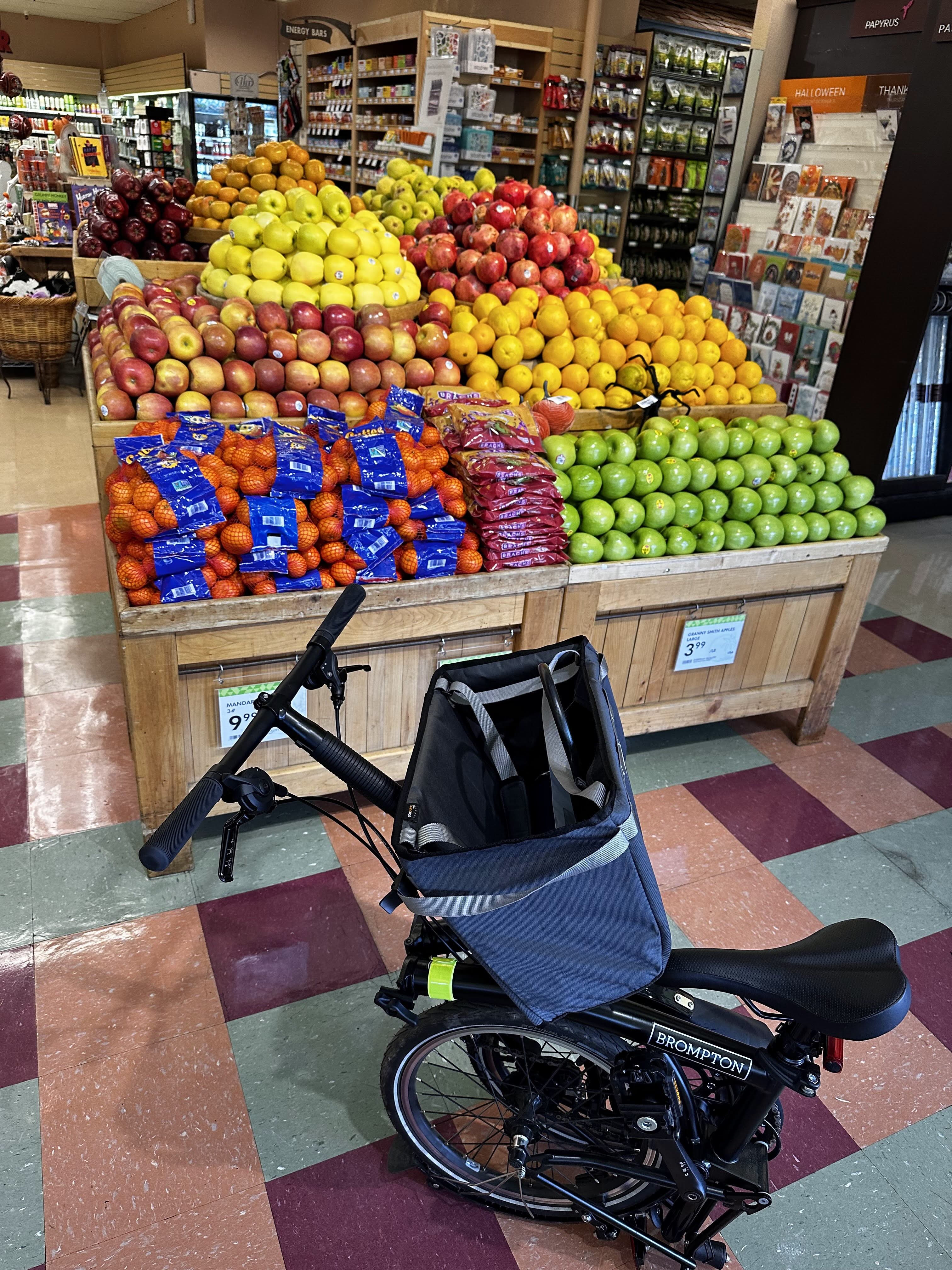 My Brompton in &lsquo;Shopping Cart Mode&rsquo; in front of a grocery fruit display. It is partially folded with the Borough basket bag mounted to the front.