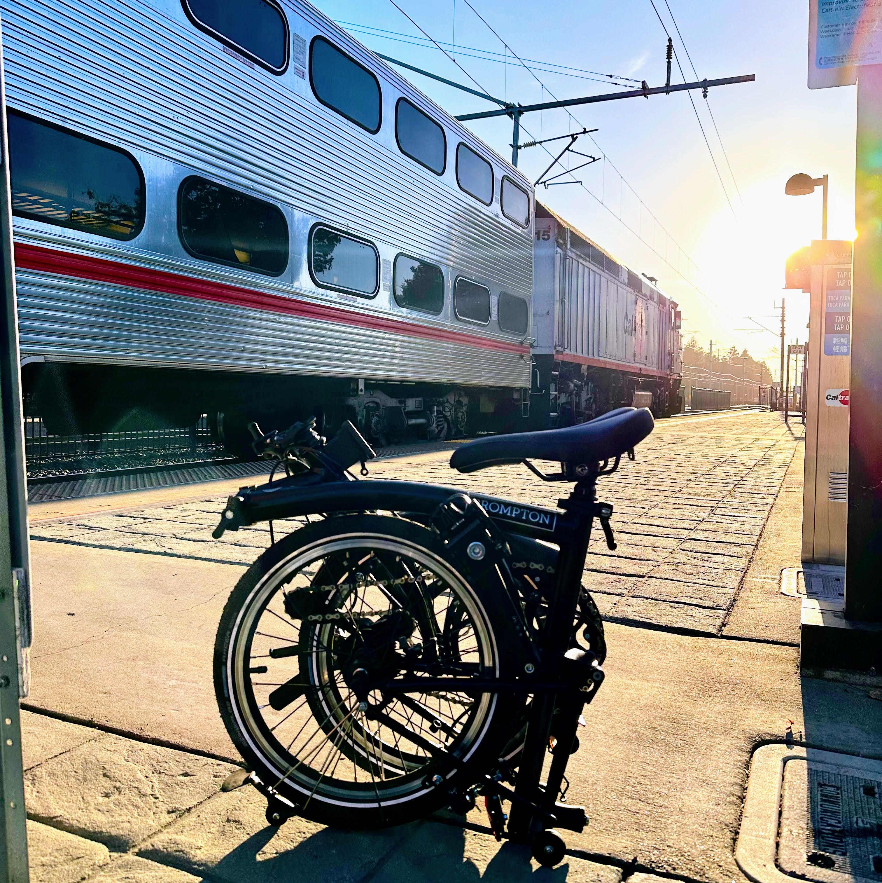 A Black Brompton C-Line bicycle fully folded. Caltrain in the background. Sun in the distance.